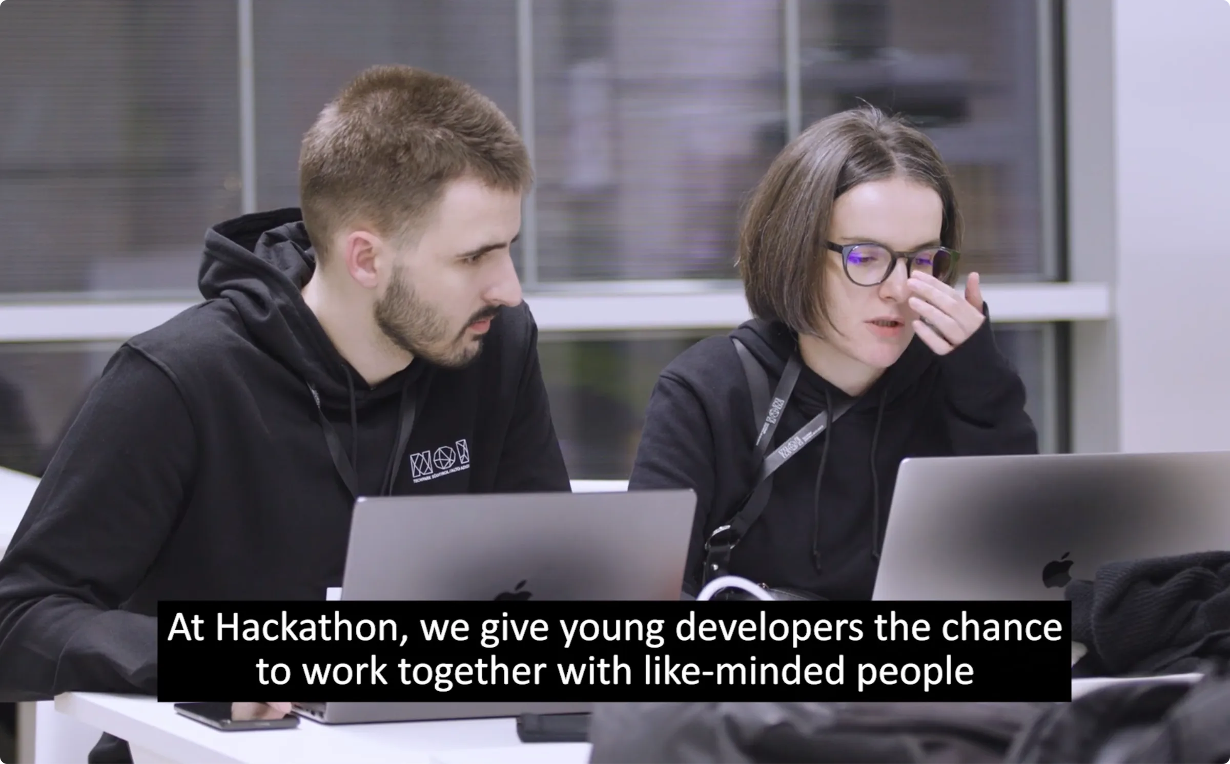 Two developers from the RouteBOT team working together on laptops at NOI Hackathon 2024, with an on-screen caption reading 'At Hackathon, we give young developers the chance to work together with like-minded people'